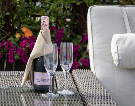 A bottle of Champagne and Champagne flutes on the outside deck on Fingal's Skerryvore deck. There are pink and orange flowers in the background. 