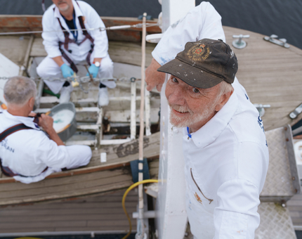 Two Yotties are sitting in a Whaler boat painting the inside. Another Yottie is looking up into the camera.