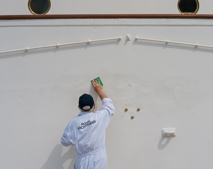 A Yottie is wiping the outside of the front of the hull with a sponge.
