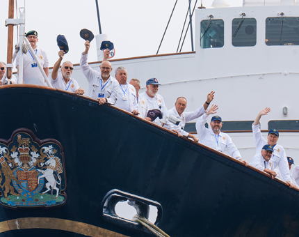 A large group of Yotties standing waving their caps in the air as they stand on the bow of The Royal Yacht Britannia. 