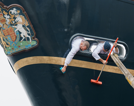 Two Yotties are reaching through a large hole on the now brushing the hull. The Royal Coat of Arms and gold line can be seen.
