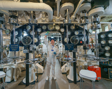 A Yottie polishing in the perfectly maintained Engine Room at Britannia. There are lots of dials and gauges and all of the chrome is gleaming.