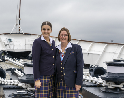 Mother and daughter stand together at the front deck of the ship, both wearing the Britannia uniform of white shirt, dark navy blazer and tartan skirt.