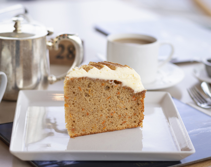 A large slice of carrot cake on a square white plate with silver teapot, and a cup and saucer in the background. 