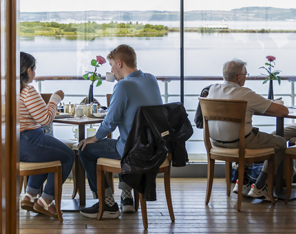 People sitting at window tables next to the water in the Royal Deck Tearoom, enjoying tea and cakes. 