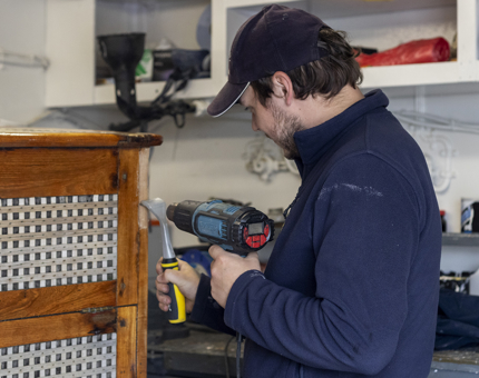 A man from the Maintenance team using a heatgun to remove varnish from a wooden box in the workshop.