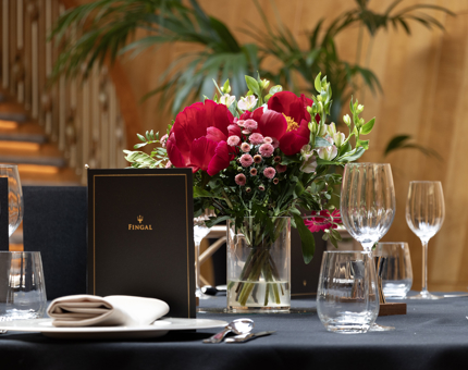 A table set-up for dinner in Fingal's Ballroom. There is bright red bouquet on the table, glasses, plates and menus. 