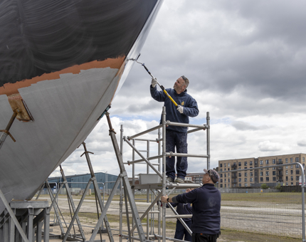 A man is standing on a small scaffold painting the hull of Bloodhound with a roller. Two other men are supporting the scaffolding. 
