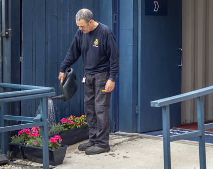 A man from the Facilities team watering a planter filled with flowers on the quayside. 
