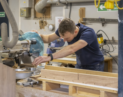 A team member from Maintenance using a saw to cut wooden handrails in the workshop.