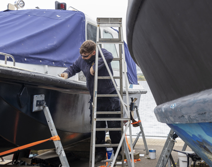 A man from the Maintenance team standing on a ladder preparing the Activity Boat for sanding. 