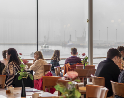 People sitting at tables in the Tearoom with a view from the windows of ships. 