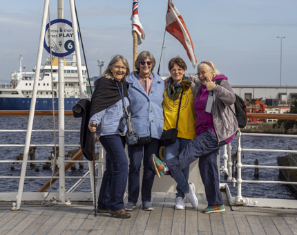A group of four ladies posing for a photo on the Verandah Deck with a ship in the background. 