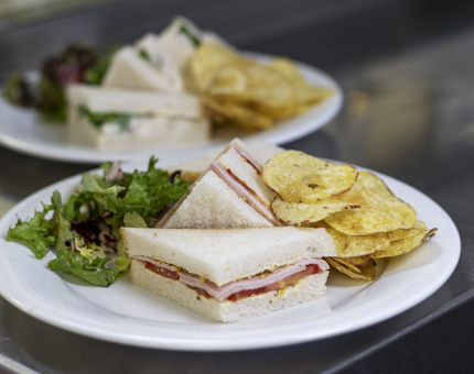 A white plate with a ham sandwich, crisps and salad on it.