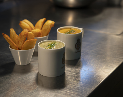 Two mugs of soup and two bowls of potato wedges sitting on a silver counter top. 