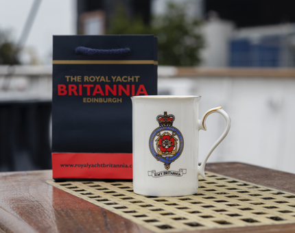 A white mug with a Britannia crest on it and a blue, red and gold gift bag sitting on a wooden bench.