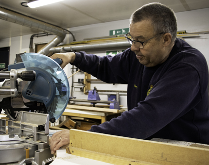 A member of the Maintenance team saws wood to manufacture a picture frame. 