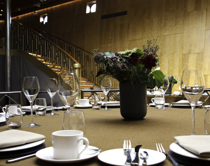 Round dining table set with plates, cutlery, cups and saucers, wine glasses and a vase of flowers in the centre. A curved staircase is in the background. 