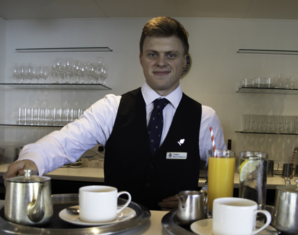 A waiter stands behind the bar with a tray containing a teapot, cups of coffee and a glass of orange juice.