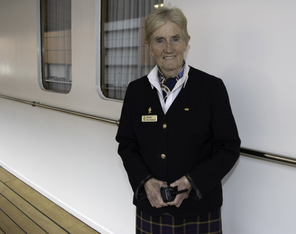 Woman in Britannia uniform smiles in greeting at visitors on Britannia's deck. 