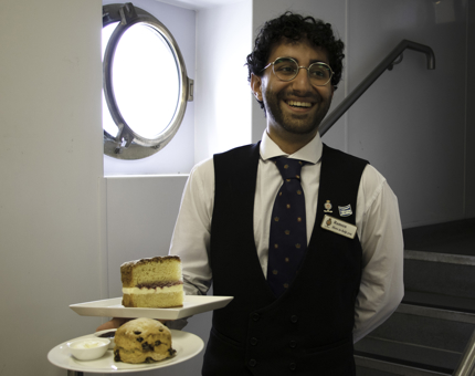 Waiter carries plates containing a fruit scone with clotted cream and jam and a slice of Victoria sponge cake. 