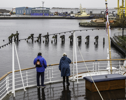 Outside Verandah Deck on a rainy day where two visitors wearing raincoats stand listening to Britannia's audio guide. 