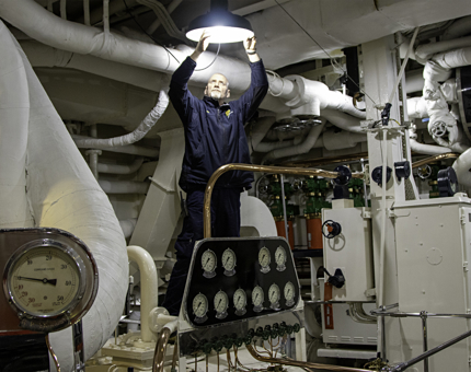 A wide shot of a maintenance team member replacing a bulb in a large overhead lamp within the Engine Room. There are many dials, pipes and machinery. 