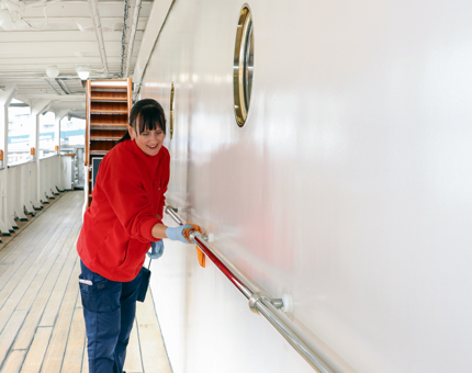 A member of the Housekeeping team wearing a red fleece polishing handrails on an outside deck. 
