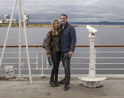 A man and a woman posting for a photo on the Verandah Deck, there is a view of the water, block of flats and hills in the background. 