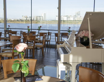 A view of the Royal Deck Tearoom with a white grad piano, tables and views over the waterfront from the large picture windows. 