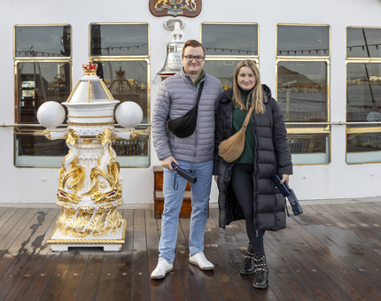 A man and a woman pose by the compass binnacle on The Royal Yacht Britannia's Verandah Deck.