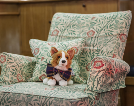 A corgi plush toy with a tartan bowtie sitting on a chair with a floral cover. 
