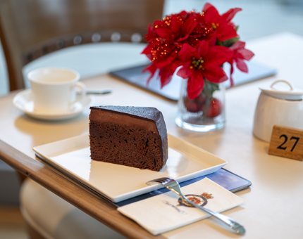 A slice of chocolate fudge cake on a table. There is a vase with red flowers in it.
