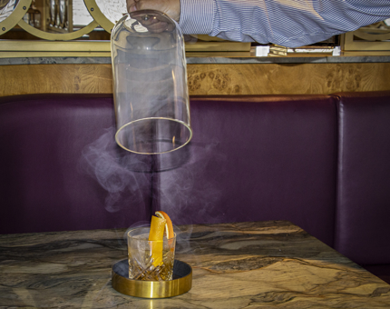 A Negroni cocktail is revealed from underneath a smoker. 
