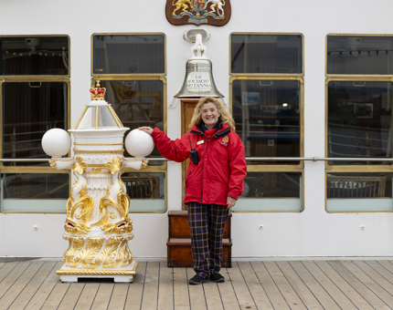A Visitor Assistant wearing a red jacket poses next to Britannia's Bell and binnacle on the Verandah Deck. 