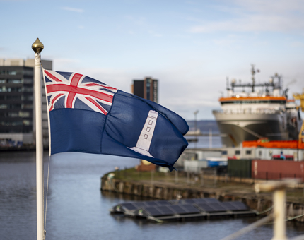 A flag with a union jack and an image of a lighthouse. There is a port in the background with ships docked.