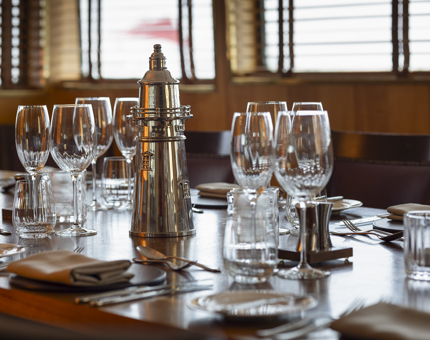 The table set in the Bridge for a meal. There are wine glasses at each setting and a silver lighthouse ornament in the middle of the table.