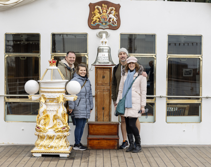 A group of four visitors posing for a photo on Britannia's Verandah Deck. They are standing next to the silver bell and white and gold binnacle. 