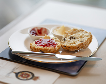 A fruit scone on a plate that has been cut open. Red jam had been spread on one side. On the plate there is also a silver knife, a pot of jam and a pot of cream. 