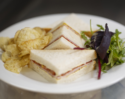 A white plate with a ham sandwich made with white bread. There is also crisps and salad on the plate. 