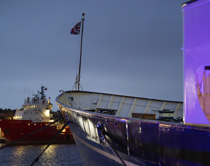 A view of the bow of Britannia looking forward in to the port. There is a ship in the background. It is dusk and the ship is lit up with a blue light. 