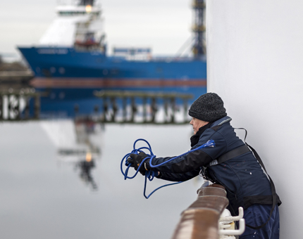 A man catching a blue rope that has been thrown up to him from a lower deck. There is a blue ship in the background. 