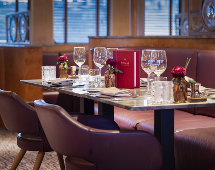 Fingal Hotel's Lighthouse Restaurant & Bar's table set for dinner. There are wine glasses and a red menu. Three small brown clear vases with dark red flowers decorate the table. The seats are a dark purple coloured leather. 