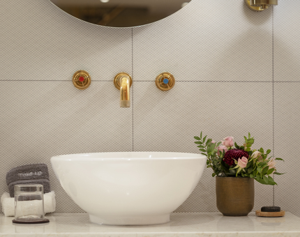 A close-up of a white sink against a white tiled wall. There is a brass tap and handles. There is a vase with pink flowers, small towels and a glass.