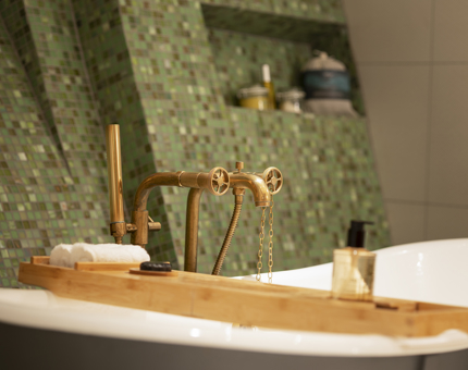 A bathroom in a Fingal Hotel cabin. The wall have small green tiles on it, there is a bath with brass taps and knobs.