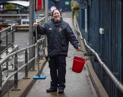 A Facilities Officer poses for a photo with a red bucket and blue brush. He is standing on a walkway next to a blue wooden fence. 
