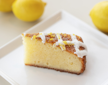 A slice of lemon drizzle cake on a white plate. There are three lemons in the background.