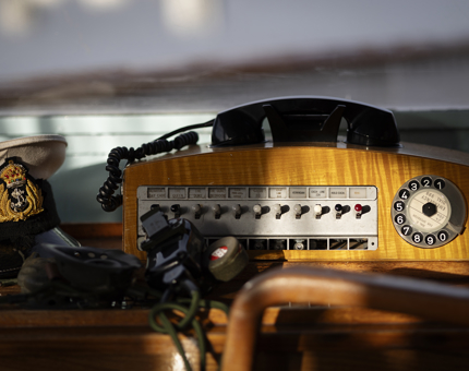 A rotary phone in the Bridge. It is made of wood and has a switch board. There is a Captain's hat sitting next to it. 