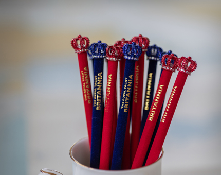 A mug with blue and red Britannia pencils. They have gold writing. The ends of the pencils feature a crown with small diamantes on it. 