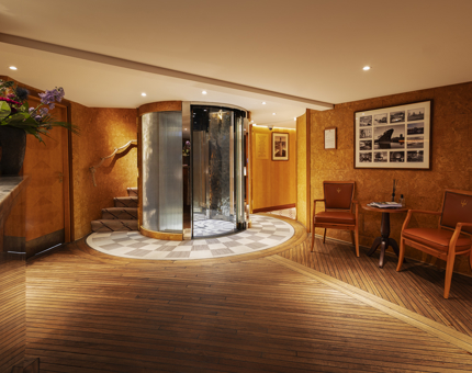The Reception area of Fingal Hotel. There is a glass circular lift on the back wall with a staircase curving behind it. There are two chairs against the righthand wall. 